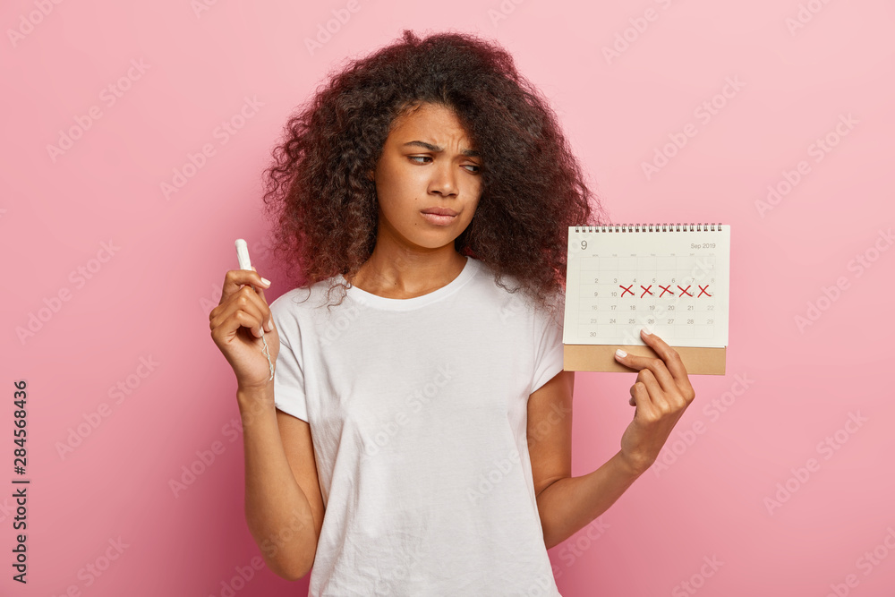 Displeased stressful dark skinned woman looks at periods calendar with marked red crosses, has painful menstruation, holds tampon for good protection. People, women health and hygiene concept Displeased stressful dark skinned woman looks at periods calendar with marked red crosses, has painful menstruation, holds tampon for good protection. People, women health and hygiene concept