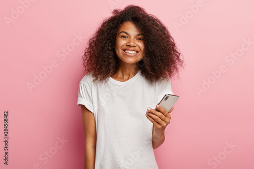 Impressed lovely Afro woman with luxurious curly hair, holds modern mobile phone, waits for call, enjoys online communication, smiles broadly, shows perfect even teeth, wears everyday t shirt