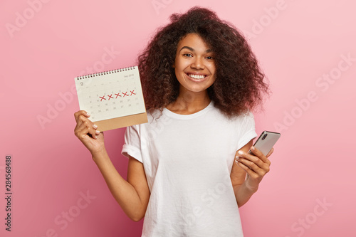 Happy curly woman holds periods calendar, checks menstruation days on mobile phone application, cares about women health, dressed casually, isolated on pink studio wall. Pms and wellness concept photo