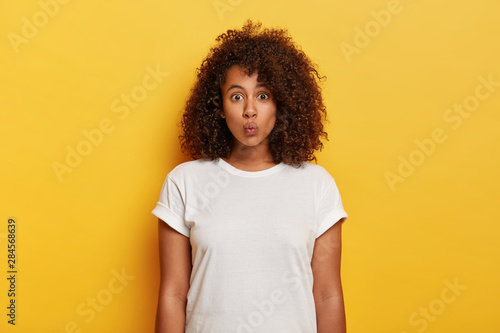 Horizontal shot of attractive flirty woman makes lips folded, waits for passionate kiss from boyfriend, has lovely outlook, curly hairstyle, wears white t shirt, models against yellow background.