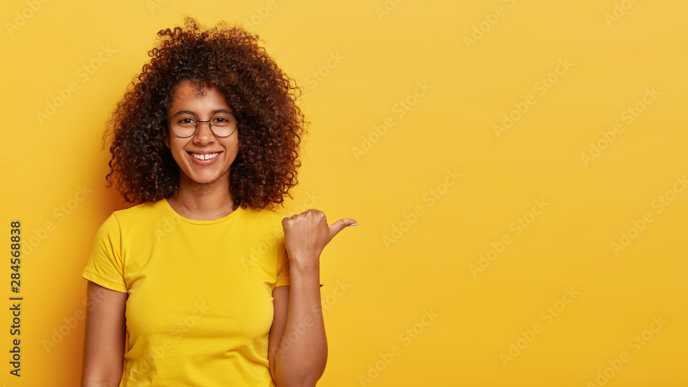 Satisfied dark skinned hipster with curly bushy hair, points away on copy space, advertises cool promo, wears transparent round eyewear and vivid casual t shirt, isolated on yellow background Satisfied dark skinned hipster with curly bushy hair, points away on copy space, advertises cool promo, wears transparent round eyewear and vivid casual t shirt, isolated on yellow background