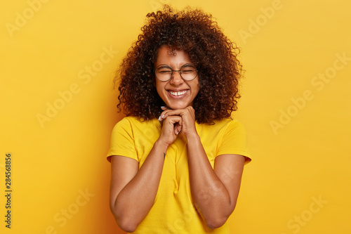 Pretty charming Afro girl with curly hair, rejoices life, keeps hands under chin, feels overjoyed and satisfied, has natural appearance, wears bright yellow t shirt, poses indoor. Happiness concept