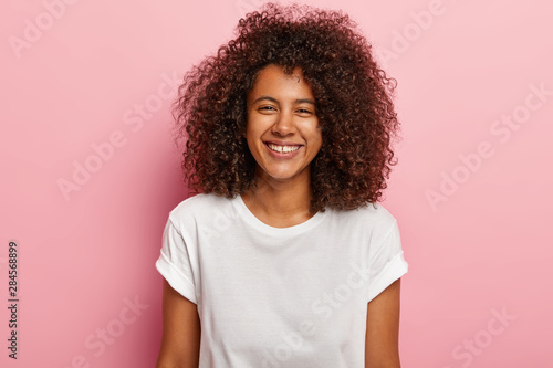 Cute happy girlfriend smiles joyfully, has white even teeth, looks at camera, enjoys great time with lover, hears funny joke, wears white t shirt, models against rosy background. Ethnicity concept © wayhome.studio 