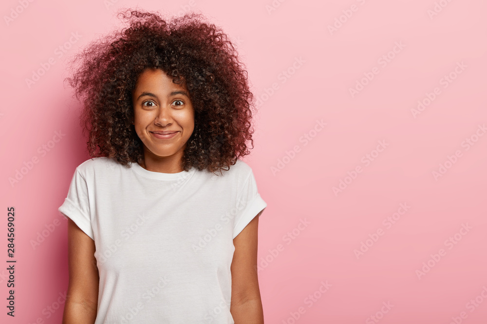 Funny good looking curly haired female teenager has pleased expression, satisfied with how things going, rejoices nice surprise or good news, wears white t shirt, models over rosy studio wall Funny good looking curly haired female teenager has pleased expression, satisfied with how things going, rejoices nice surprise or good news, wears white t shirt, models over rosy studio wall