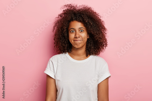 Photo of funny woman has curly thick hair, presses lips together, has happy face, wears white t shirt, isolated over pink background. Good looking young African American girl expresses happiness.