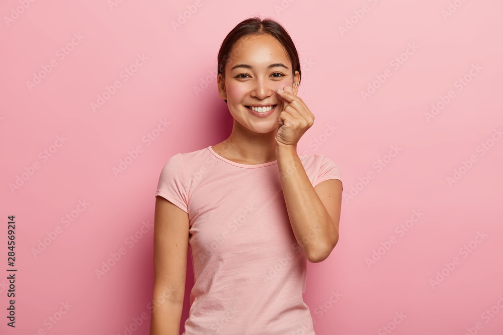 Lovely young female model touches genlty her rouge cheeks, shows healthy skin on face, has Asian appearance, smiles broadly, wears pink t shirt, poses indoor. People, ethnicity, beauty, skin care Lovely young female model touches genlty her rouge cheeks, shows healthy skin on face, has Asian appearance, smiles broadly, wears pink t shirt, poses indoor. People, ethnicity, beauty, skin care