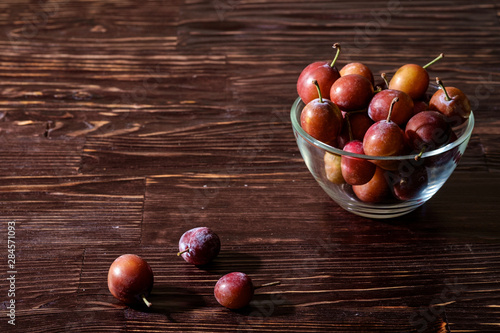 Ripe sweet plum fruits in glass bowl near with scattered plums on dark moody wood table background  hard light  copy space