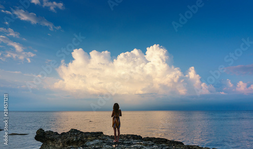 Girl on a rock, overlooking the vast calm sea in front of her, gazing at the beautiful clouds in a distance