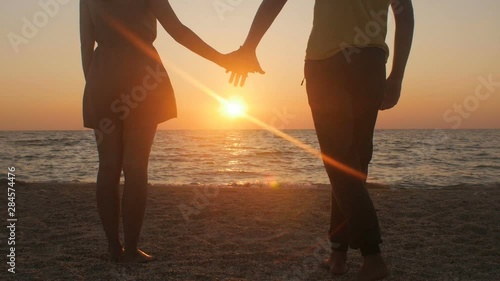 silhouette of a romantic young couple standing on the seashore, man comes up and takes the girl's hand and together enjoying sunrise on the beach, summer vacation and love