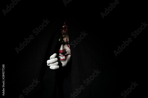 Photo A terrible scary clown in a colored wig peeps out from behind black curtains