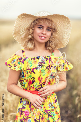 Portrait of a beautiful girl in a hat holding ripe spikelets of barley in her hands. A model in a colored sundress poses on the background of an agricultural field, close-up
