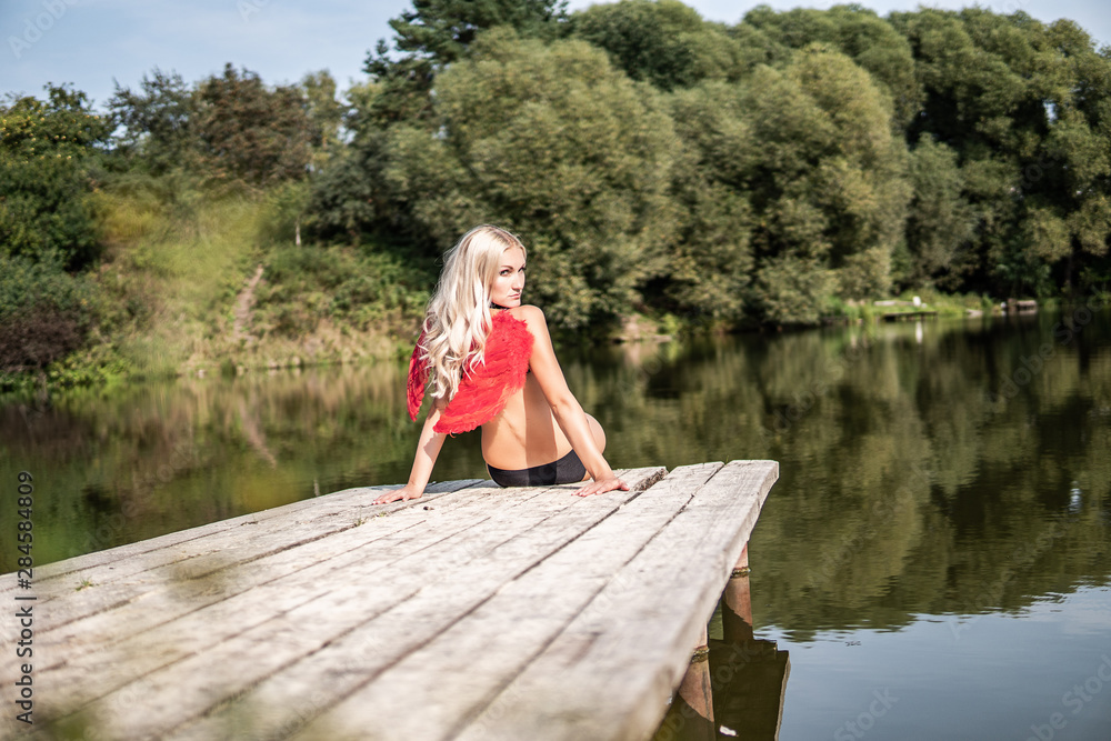 Fairy girl in Angel Red Wings sits on the quay on the river bank. gently turning head Fairy girl in Angel Red Wings sits on the quay on the river bank. gently turning head