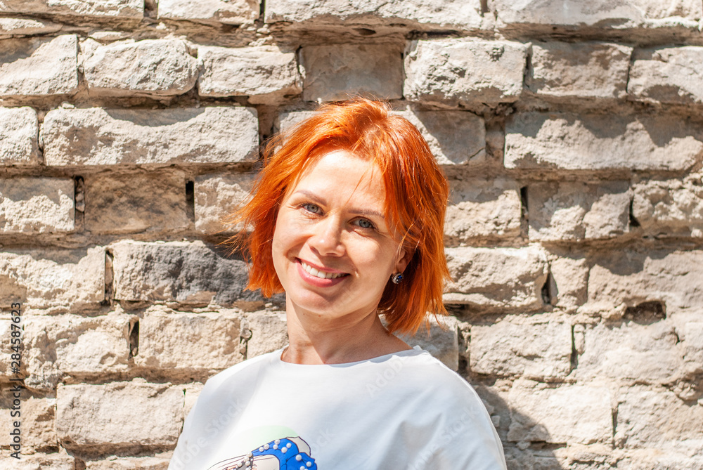 A young girl with red hair in a white T-shirt and a bright green skirt stands near the brick wall on a city street and smiles. A young girl with red hair in a white T-shirt and a bright green skirt stands near the brick wall on a city street and smiles.