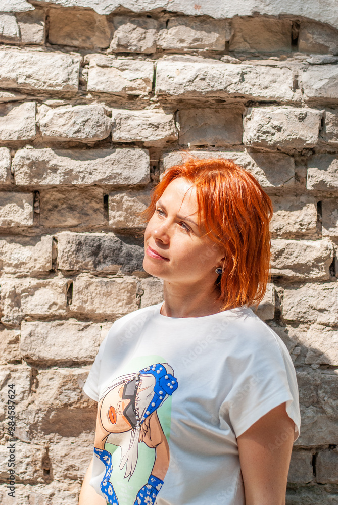 A young girl with red hair in a white T-shirt and a bright green skirt stands near the brick wall on a city street and looks to the side. A young girl with red hair in a white T-shirt and a bright green skirt stands near the brick wall on a city street and looks to the side.