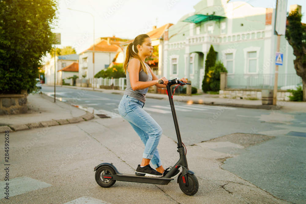 Young woman rides electric kick scooter on the street waiting on the crossroad wearing jeans in summer day evening traffic transport Young woman rides electric kick scooter on the street waiting on the crossroad wearing jeans in summer day evening traffic transport