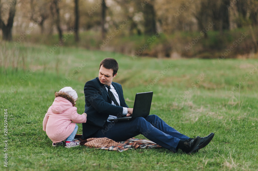 A serious businessman in a suit sits in the park on the green grass and works on a laptop, a small child walks alongside and distracts. Computer, lifestyle, work. Father and daughter, business. A serious businessman in a suit sits in the park on the green grass and works on a laptop, a small child walks alongside and distracts. Computer, lifestyle, work. Father and daughter, business.