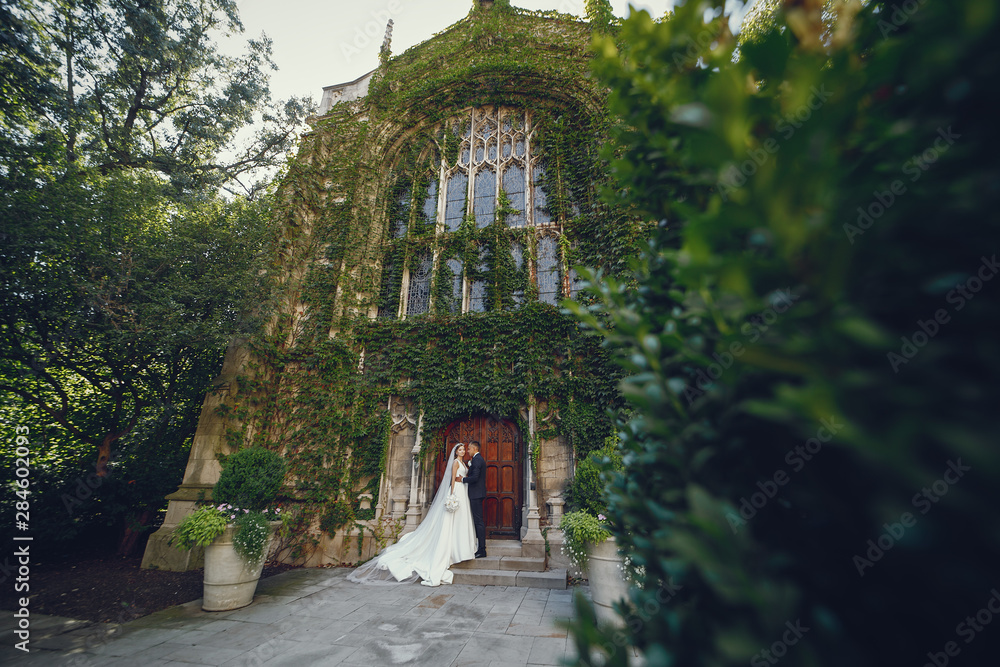 Fototapeta premium Elegant bride in a white dress and veil. Handsome groom in a blue suit. Couple near large building