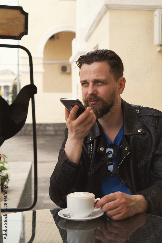Stylish hipster freelancer drinking coffee on a summer terrace of a restaurant. A man is recording a voice message. Handsome guy with a fashionable haircut and neat beard Stylish hipster freelancer drinking coffee on a summer terrace of a restaurant. A man is recording a voice message. Handsome guy with a fashionable haircut and neat beard