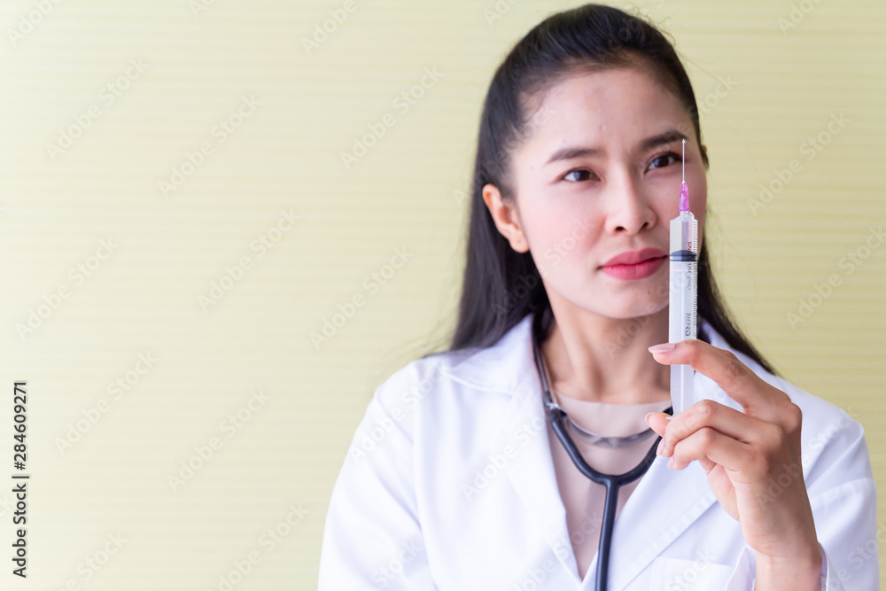 The female Asian doctor wearing a white robe sitting in a chair with a stethoscope on the neck be a serious face and preparing a syringe for patients. The female Asian doctor wearing a white robe sitting in a chair with a stethoscope on the neck be a serious face and preparing a syringe for patients.