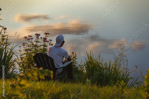 Man in the seat at bank of lake also looks afar on sunset  reflecting the shadow from the orange sky of the sunset. The film grain