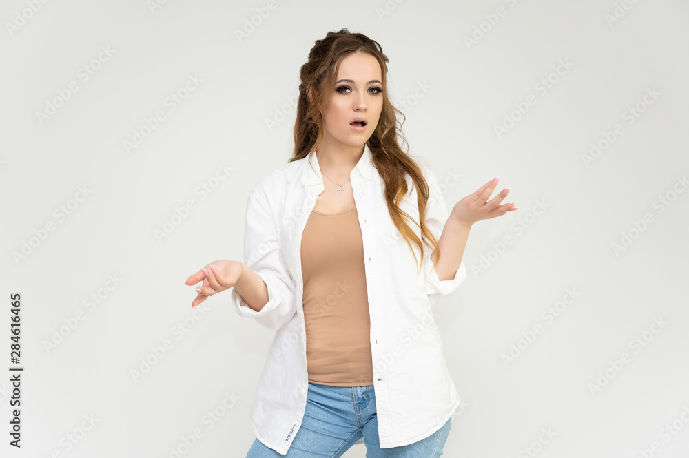 Photo portrait above the knee of a pretty brunette woman girl with long beautiful curly hair on a white background in a white shirt and blue jeans. Talking while standing in front of the camera. Photo portrait above the knee of a pretty brunette woman girl with long beautiful curly hair on a white background in a white shirt and blue jeans. Talking while standing in front of the camera.