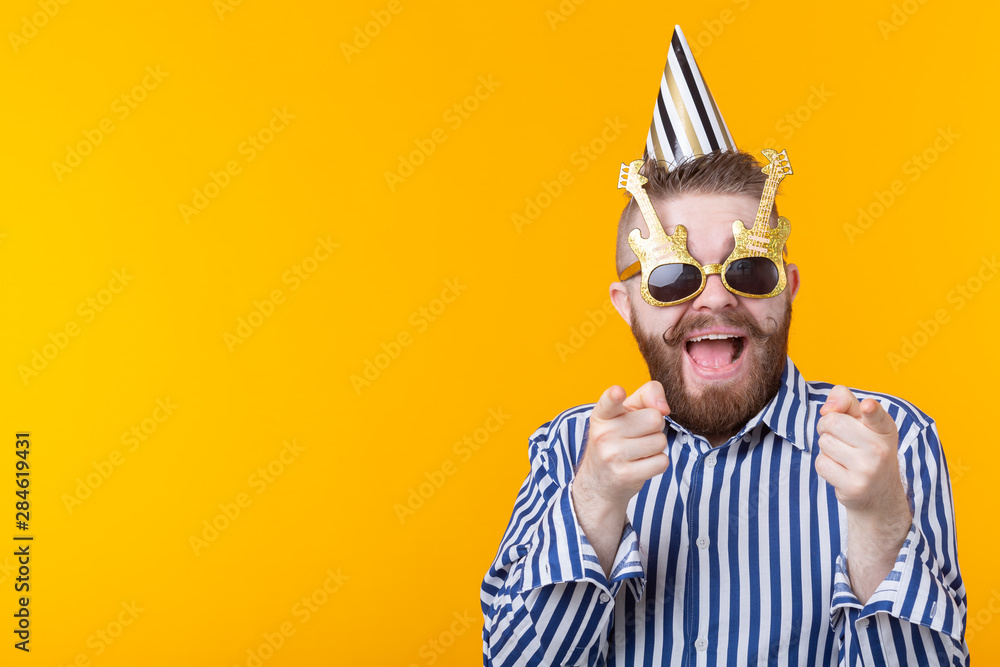 Joyful young hipster male in a paper cap and glasses is laughing happily on a yellow background with copy space. The concept of a mega party and holiday.