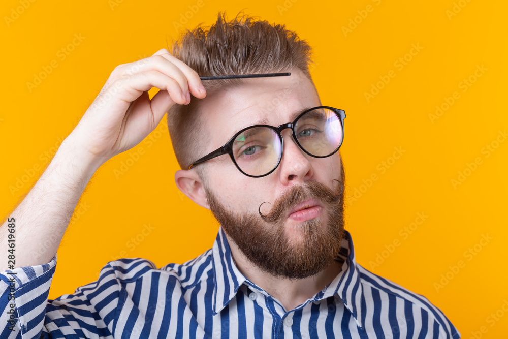 Obraz premium Portrait of a funny handsome young male hipster with a mustache and beard combing his hair against a yellow background. Barbershop concept and self-care.
