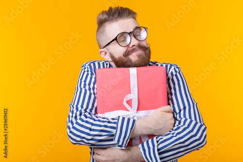 Joyful young hipster guy with a mustache in glasses hugs a red gift box on a yellow background. The concept of the joy of gifts.