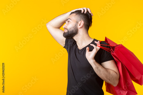 Side view of an upset young bearded stylish hipster man holding shopping bags posing on a yellow background. Concept of superfluous purchases.