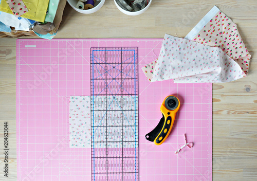 Work place: cotton fabrics, pink cutting mat, quilting ruler, rottary cutter, sewing supplies in white cups and wooden desk photo