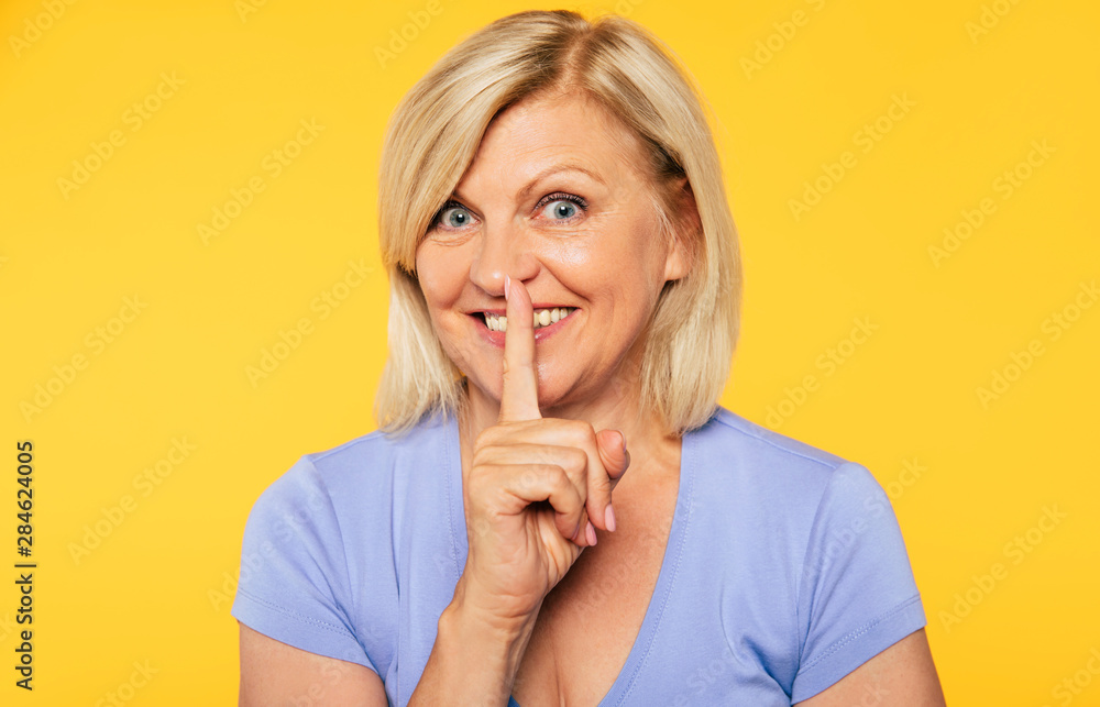 Close up photo of beautiful blonde senior woman while she holds finger on her lips and shows silence gesture on yellow wall Close up photo of beautiful blonde senior woman while she holds finger on her lips and shows silence gesture on yellow wall