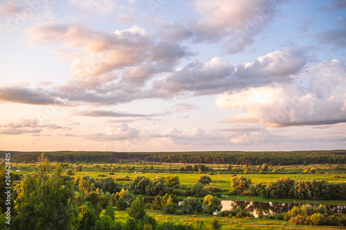 Great amazing sunset countryside landscape. Aerial view of green valleys, trees, river water and perfect golden and blue cloudscape over beautiful summer nature. Horizontal color photography.