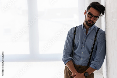 Portrait of handsome bearded hipster guy with glasses on standing indoors by the white brick wall © lordn