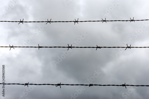 Barbed wire background. Three horizontal lines of barbed wire against a dramatic dark cloudy sky in monochrome colors. No access, trespassing or captivity symbol.