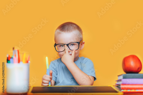 Happy blonde clever boy is sitting at a desk in glasses and smiling. Ready for school. Back to school. Apple, pens and books on desk