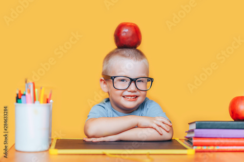 Happy cute clever boy is sitting at desk and smiling. Child is on a yellow background. First time to school. Back to school. pupil in glasses with apple on his head