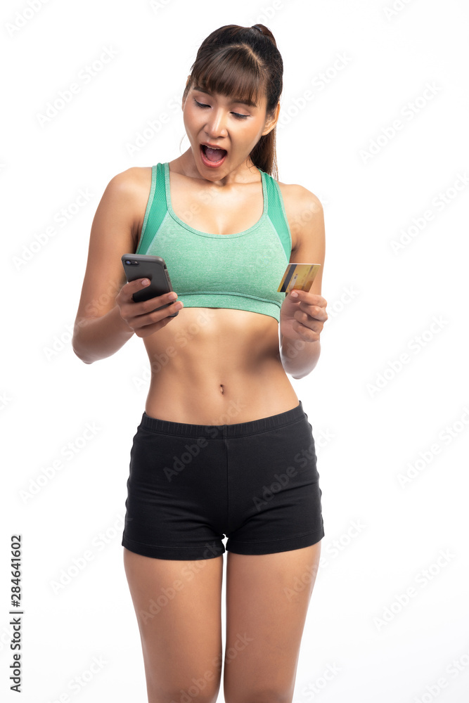Fitness woman using mobile phone and holding credit card isolate in white background. Asian girl, extremely surprise look.
