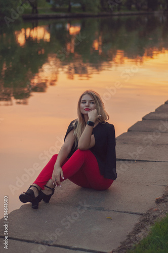 The girl in red trousers sits on the embankment of the river at sunset © Yekatseryna
