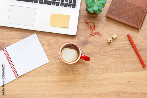 Wooden office desk table with notebook, laptop, glasses, plant, red cup of cappuccino and other office supplies. Top view with copy space, flat lay.