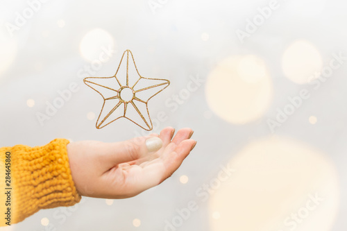 Golden Christmas star levitating above female hand in orange sweater, garland bokeh on background