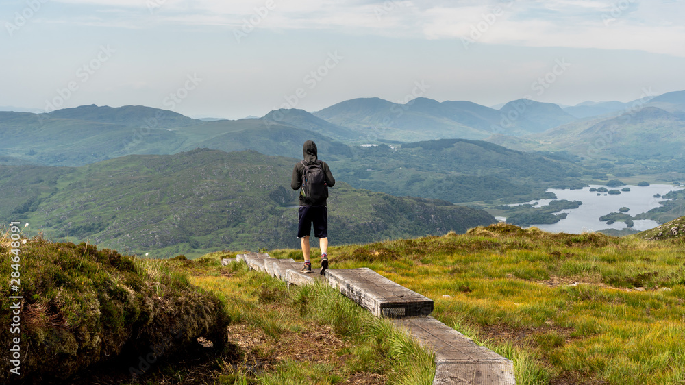 Male hiker walking on a boardwalk trail through bogland and heathland in the Irish mountains on a hazy summer day. Hiking in a scenic landscape on Torc Mountain in Ireland.