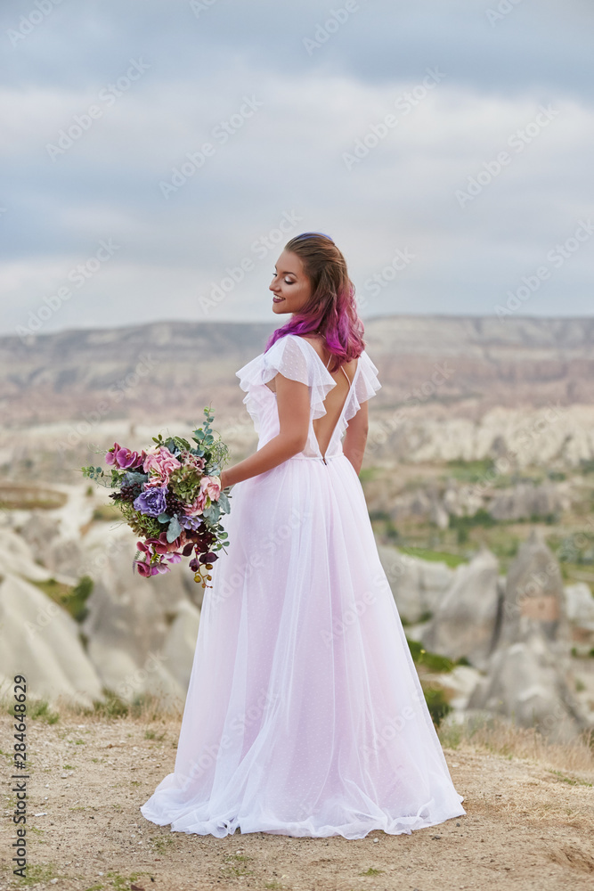 Woman with beautiful bouquet of flowers in hands stands on mountain in rays of dawn sunset. Beautiful white long dress on the girl body. Perfect bride with pink hair. Turkey, Cappadocia