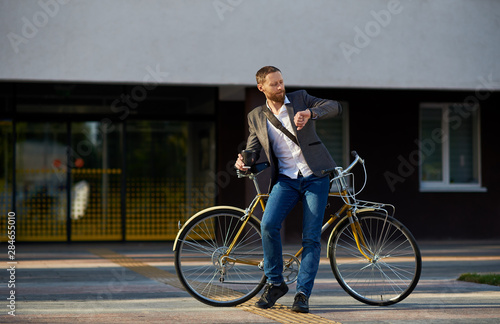 A young stylish bearded  businessman in suit going to work by bike. Drinking coffee from a cup to go and talking on a mobile  phone on the background of an office building. Takes a working break © oes