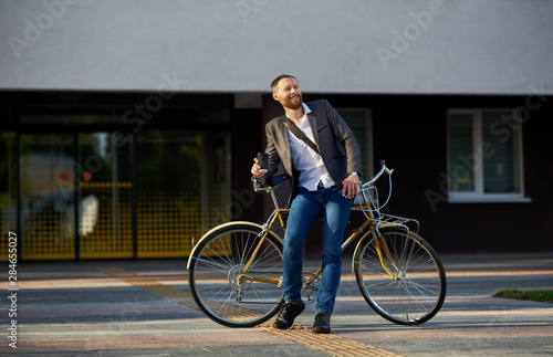 Fototapeta Naklejka Na Ścianę i Meble -  A young stylish bearded  businessman in suit going to work by bike. Drinking coffee from a cup to go and talking on a mobile  phone on the background of an office building. Takes a working break