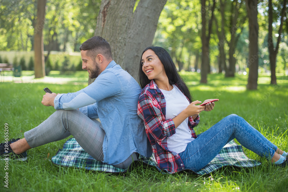 loving couple sitting on a plaid blanket in the park and use the phone, handsome guy and girl loving couple sitting on a plaid blanket in the park and use the phone, handsome guy and girl