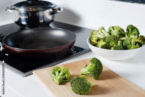 Pile of green raw uncooked broccoli in white ceramic bowl on countertop with empty frying pan on electric induction stove. Vegan diet concept. Close up, copy space, background, top view.