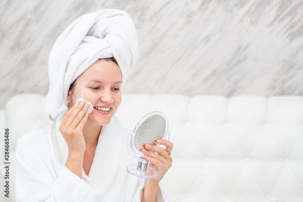 Beautiful smiling young woman applies a tonic on her face or removes makeup in front of a mirror at home Beautiful smiling young woman applies a tonic on her face or removes makeup in front of a mirror at home