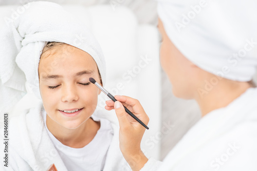 Happy family at home. Mother and little daughter while applying makeup at home. Mom and child girl are in bathrobes and with towels on their heads