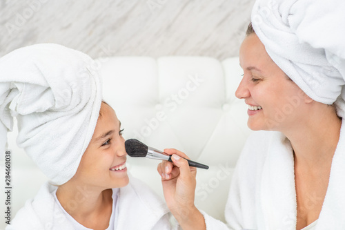 Smiling mother applying powder on nose her little daughter using a brush. Mom and child girl are in bathrobes and with towels on their heads