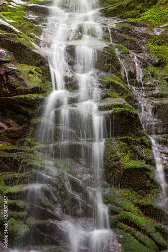 Impressive, scenic mountain waterfall cascading down the rocky cliff covered by colorful, vivid green moss and patches of sun hitting the rocks